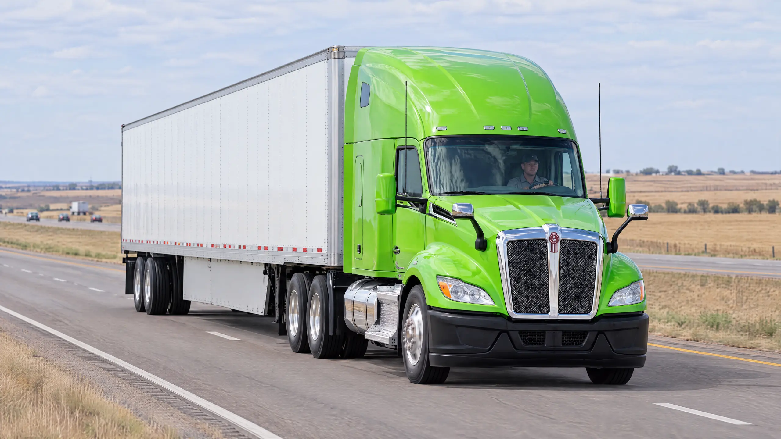 A green semi tractor pulling a white 53-foot dry van down a divided highway through open farmland