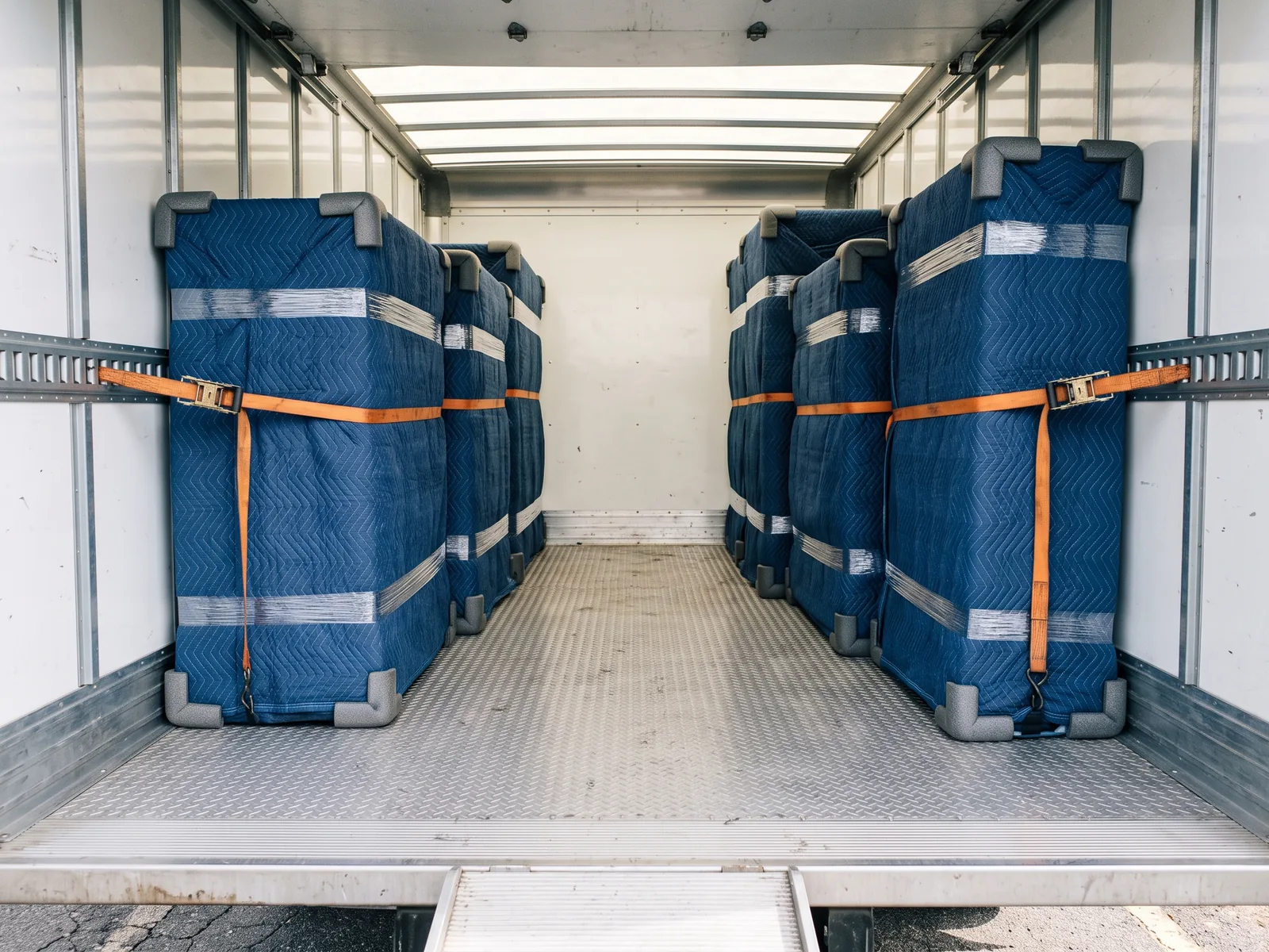 Blanket-wrapped freight standing corner-protected inside a box truck, held against the E-track wall rails by orange ratchet straps on a diamond-plate metal floor